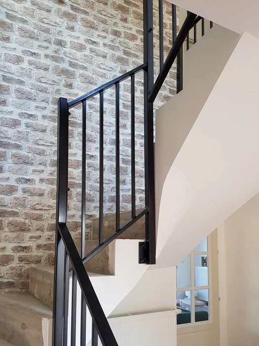 Espace de vie moderne avec escalier métallique noir, mur en briques blanches peintes, bureau sous escalier avec chaise ergonomique, canapé gris, table basse design